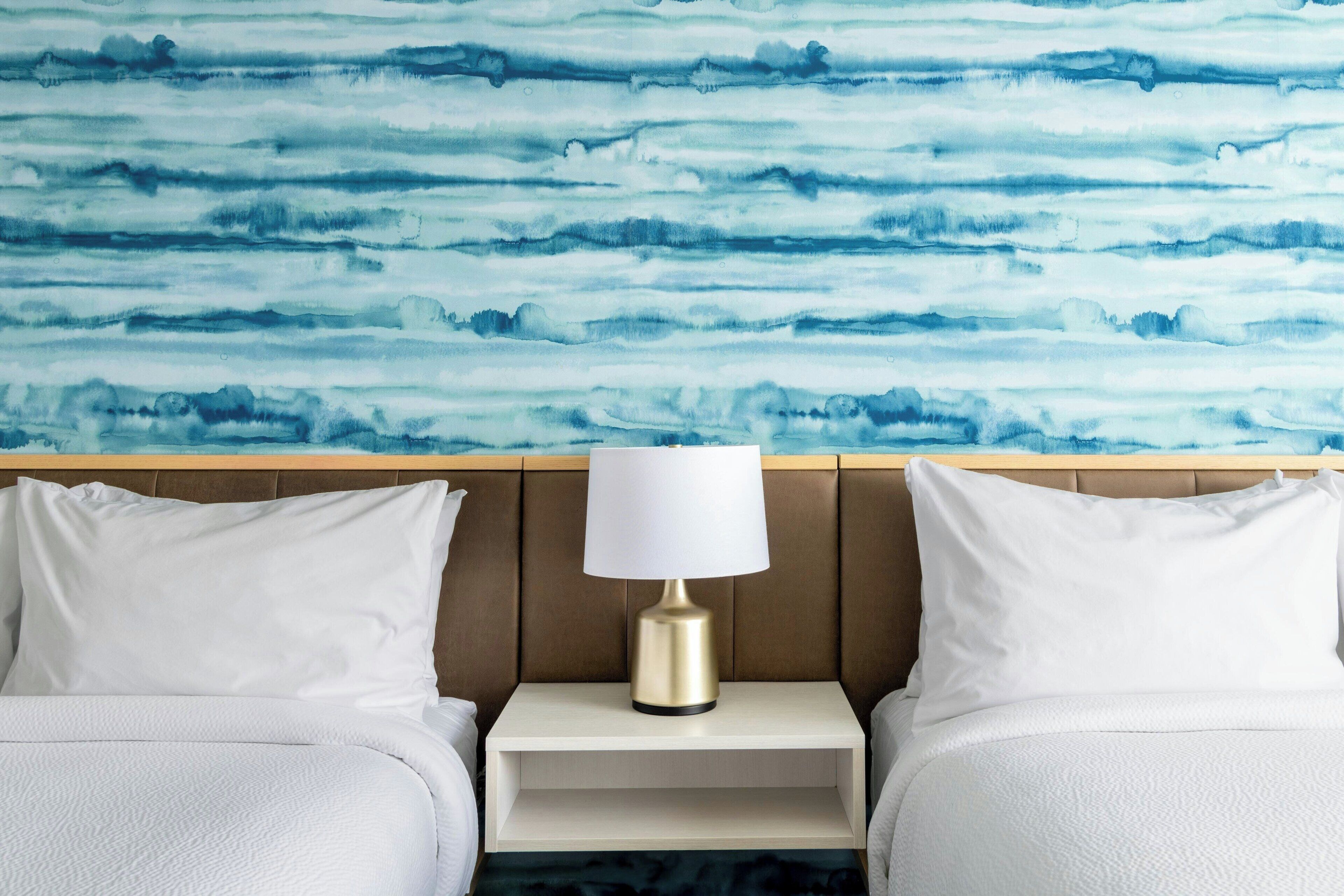 A hotel room with a bed, white pillows, a bedside table with a lamp, and a wall with a blue abstract pattern.