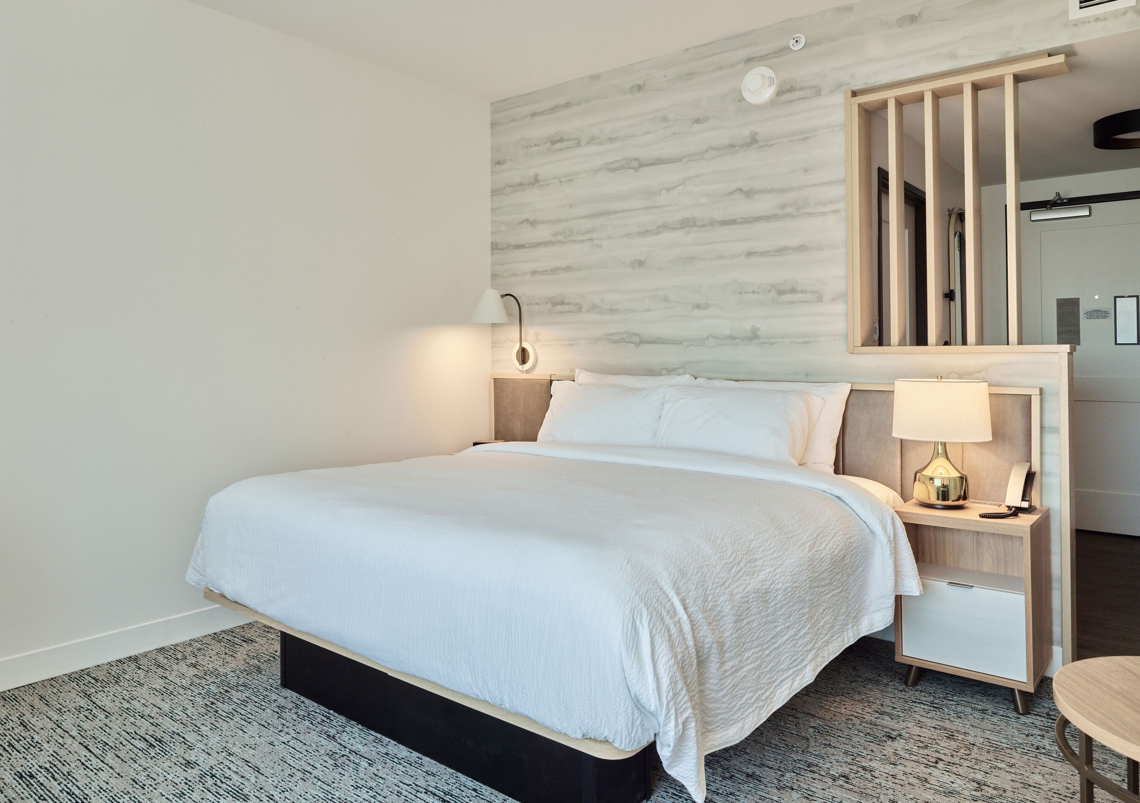 A modern hotel room with a large bed, a nightstand with a lamp, a mirror, and a small table.