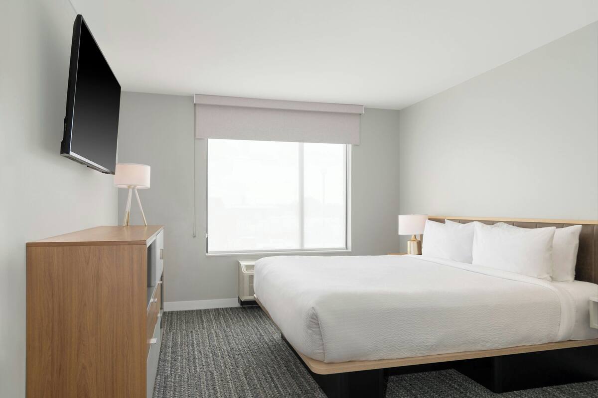 Habitación de hotel con cama, mesitas de noche, televisor de pantalla plana montado en la pared, ventana con persiana clara y radiador.