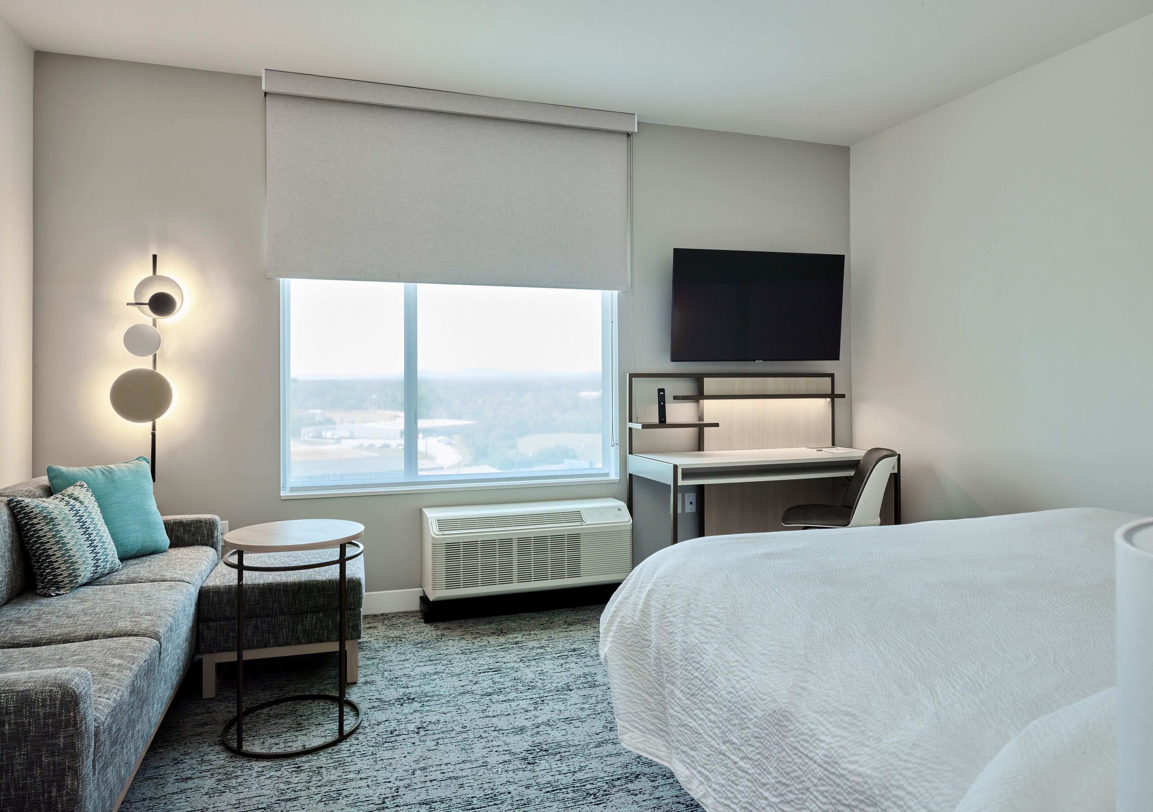 A hotel room with a bed, a sofa, a desk, a flat-screen TV, a window with a view, and a wall-mounted air conditioner.