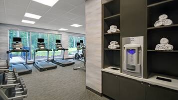 Et fitnesscenter med løbebånd, crosstrainere og styrkemaskiner. Der er vinduer med udsigt til grønne områder, og en drikkestation med håndklæder på hylder.