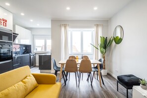 Una cocina moderna con electrodomésticos integrados, una zona de comedor con mesa y sillas, un sofá amarillo y una planta grande en maceta.