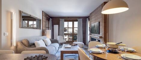 A modern living room with a sofa, dining table, and chairs. There is a fireplace and a television mounted on the wall.