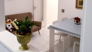 Uma sala de jantar com mesa e cadeiras brancas, uma planta verde no balcão, um sofá branco e uma porta que dá para outro cômodo.