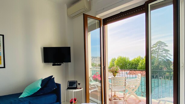Quarto com cama, sofá azul, televisão e varanda com mobiliário de exterior.