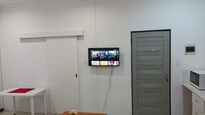 A room with a television mounted on the wall, a wooden table with a potted plant, a white door, and a small cabinet with a microwave. - Danodeb Lodge  (Pietermaritzburg)