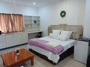 A bedroom with a bed, a desk, a chair, a small table, a refrigerator, and a shelf unit.