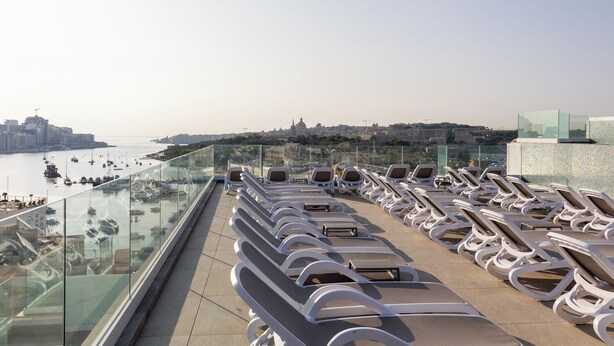 Terraço no topo do edifício com espreguiçadeiras e guarda-corpo de vidro, com vista para um porto e o horizonte da cidade.