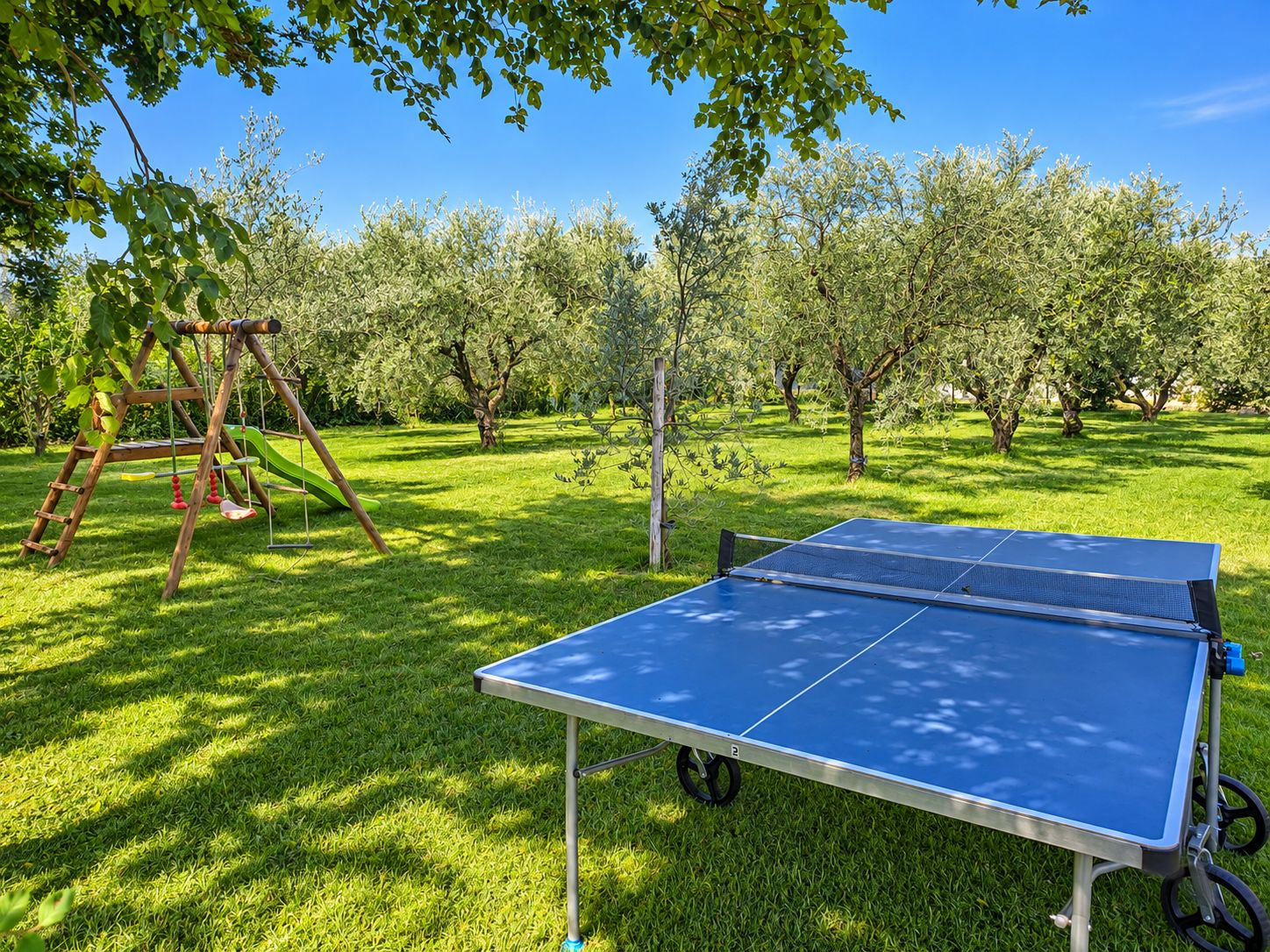 A ping pong table in a grassy outdoor area with a swing set and trees in the background.