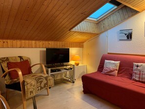 Quarto de loft aconchegante com teto inclinado, televisão, sofá vermelho e poltrona estampada.