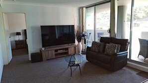 En stue med et fladskærms-tv på et træstel, et glas sofabord, en lædersofa med mønstrede puder og et stort vindue med udsigt over landskabet.