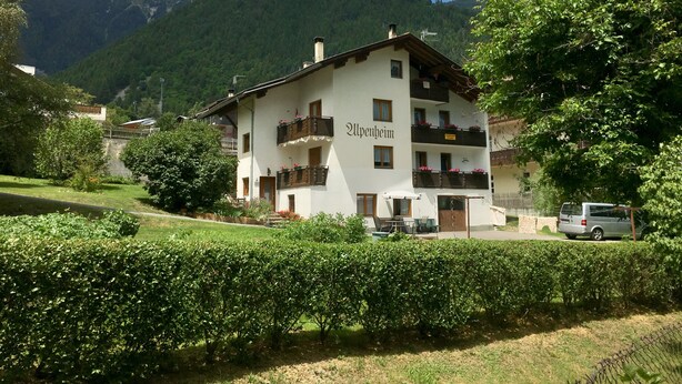 A multi-story building with balconies, named 'Alpenheim', surrounded by lush greenery and mountains in the background.