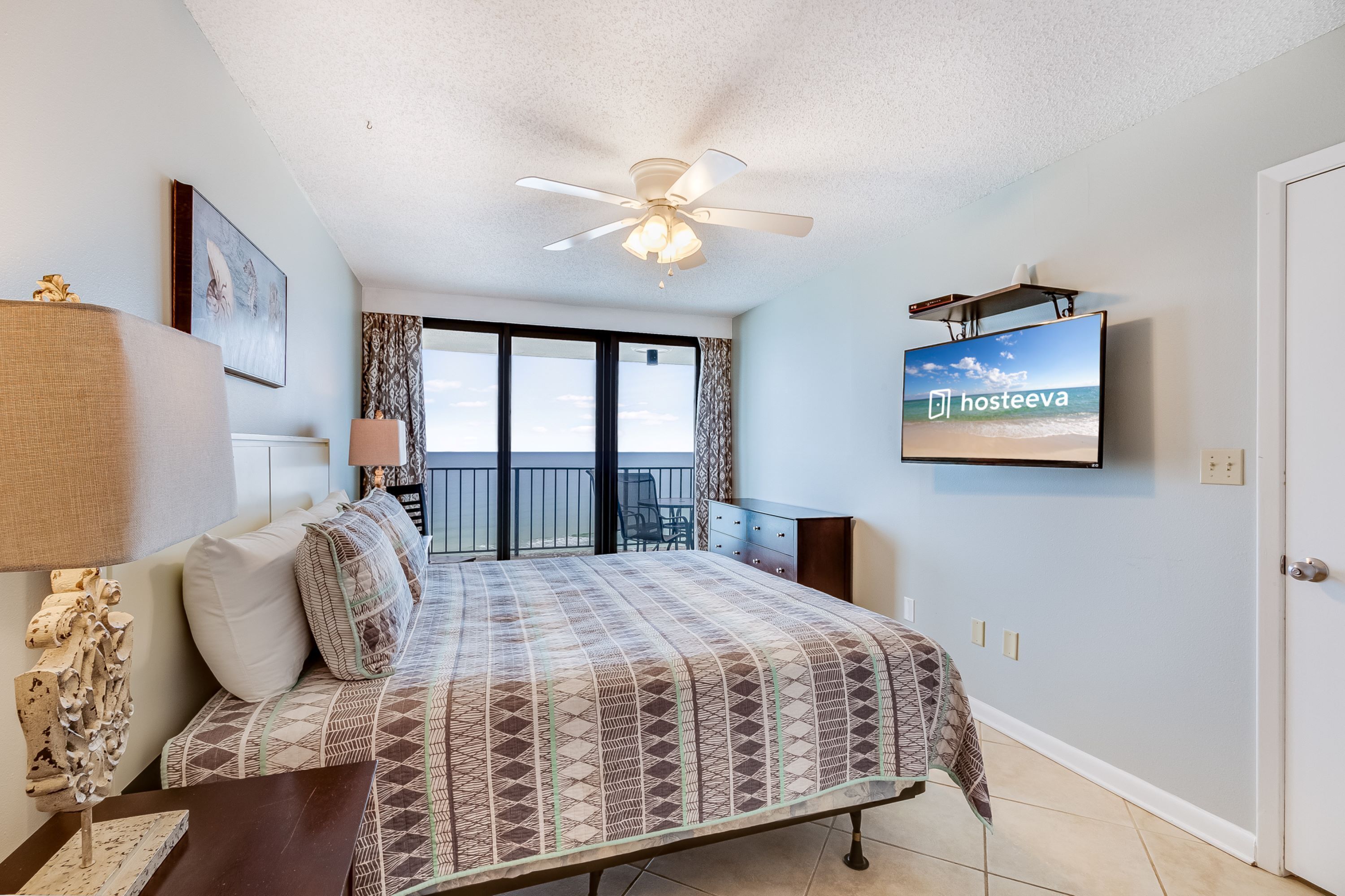 A bedroom with a bed, a ceiling fan, a TV on the wall, and a view of the sea through the window.