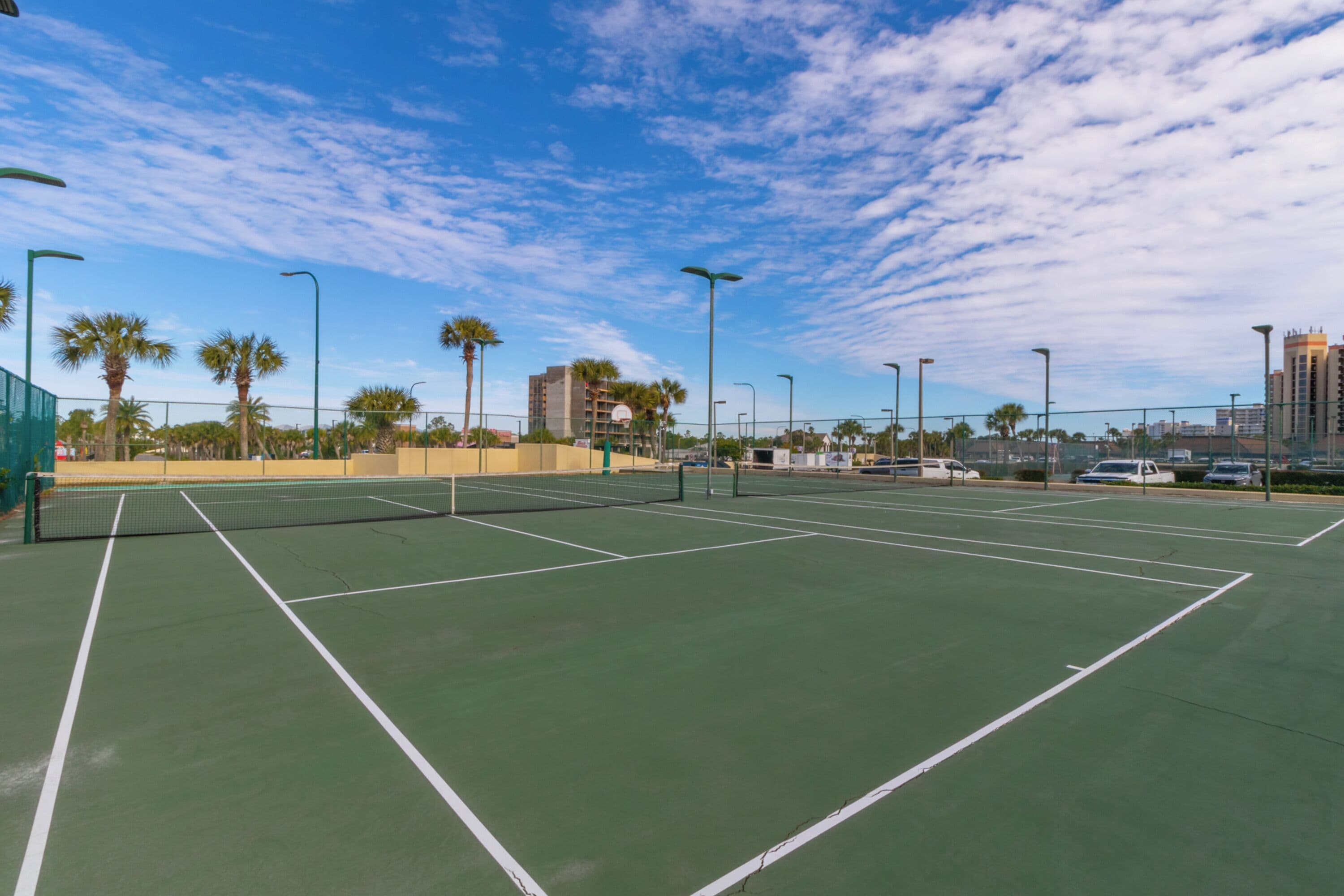 Cancha de tenis exterior con varias canchas, rodeada de palmeras y farolas.