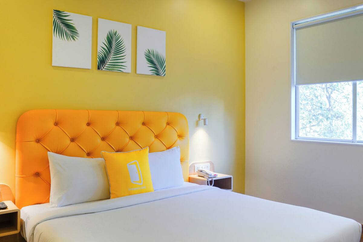 Une chambre à coucher avec une tête de lit orange à motifs capitonnés, une literie blanche et trois œuvres d’art murales ornées de motifs de feuilles.