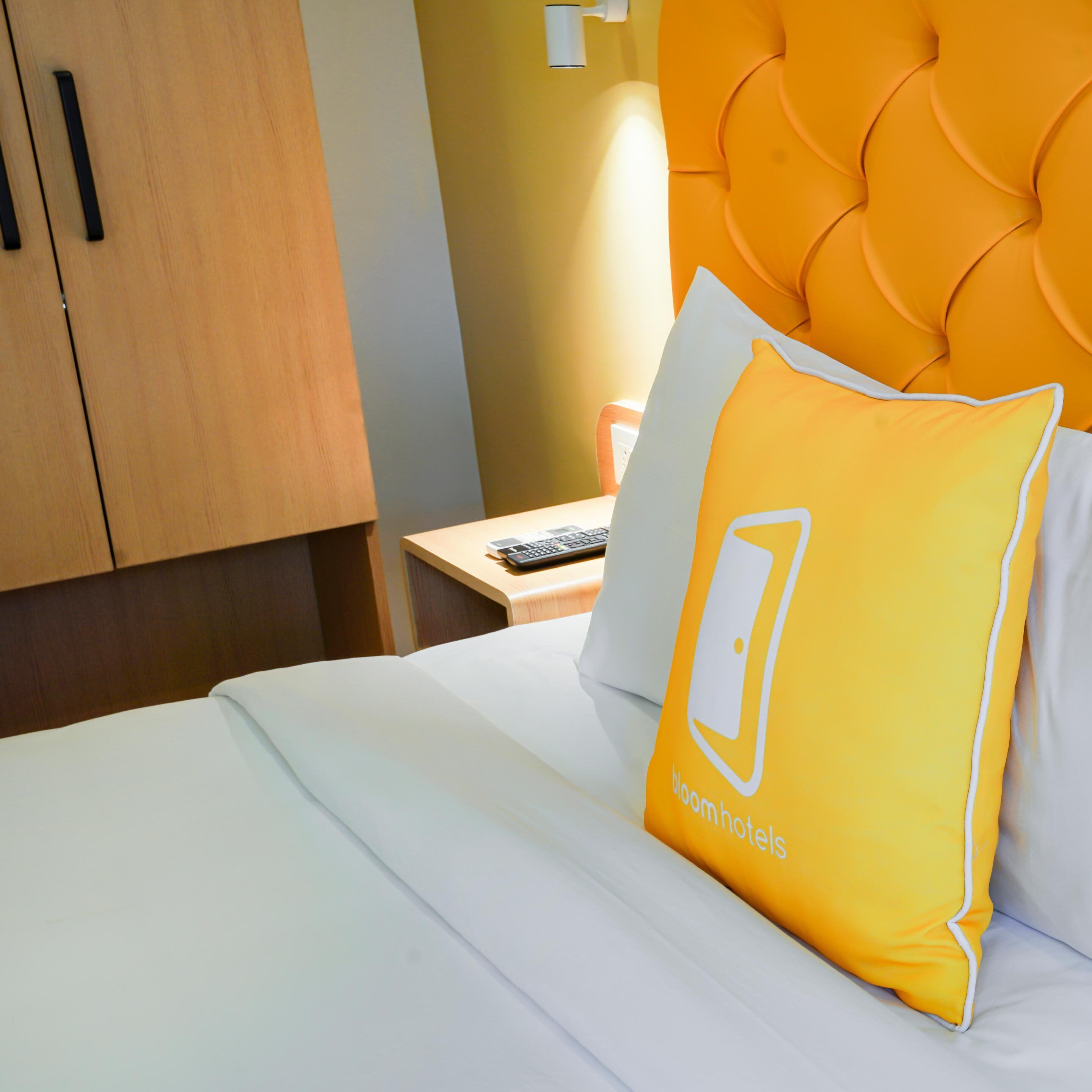 A hotel room with a bed, a wooden wardrobe, a bedside table with a remote control, and a bright orange headboard.