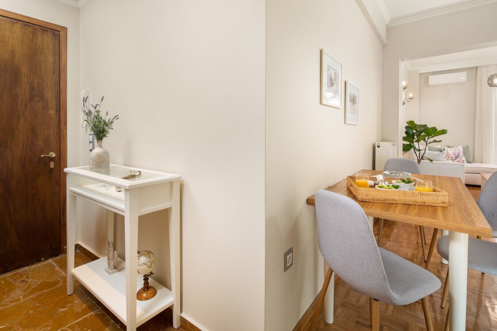 Ein Zimmer mit Holztür, einem weißen Konsolentisch mit Globus und Dekoration, einem Esstisch mit Stühlen und einem Frühstückswagen, sowie einer weißen Wand mit gerahmten Bildern.