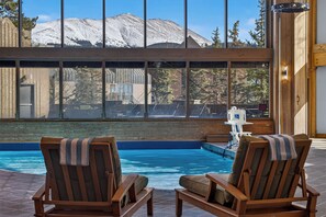 A pool area with two wooden lounge chairs, a view of a mountain, and a building with a glass facade.