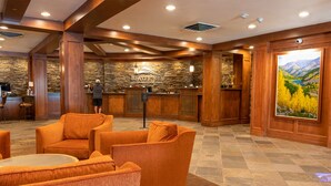 Un hall d’entrée avec des boiseries, un mur en pierre et un grand tableau représentant un paysage de montagne. On y trouve des fauteuils recouverts de tissu orange et un comptoir d’accueil où se tient une personne.