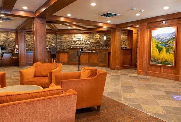 A lobby with wooden paneling, a stone wall, and a large painting of a mountain landscape. There are orange upholstered chairs and a reception desk with a person behind it.