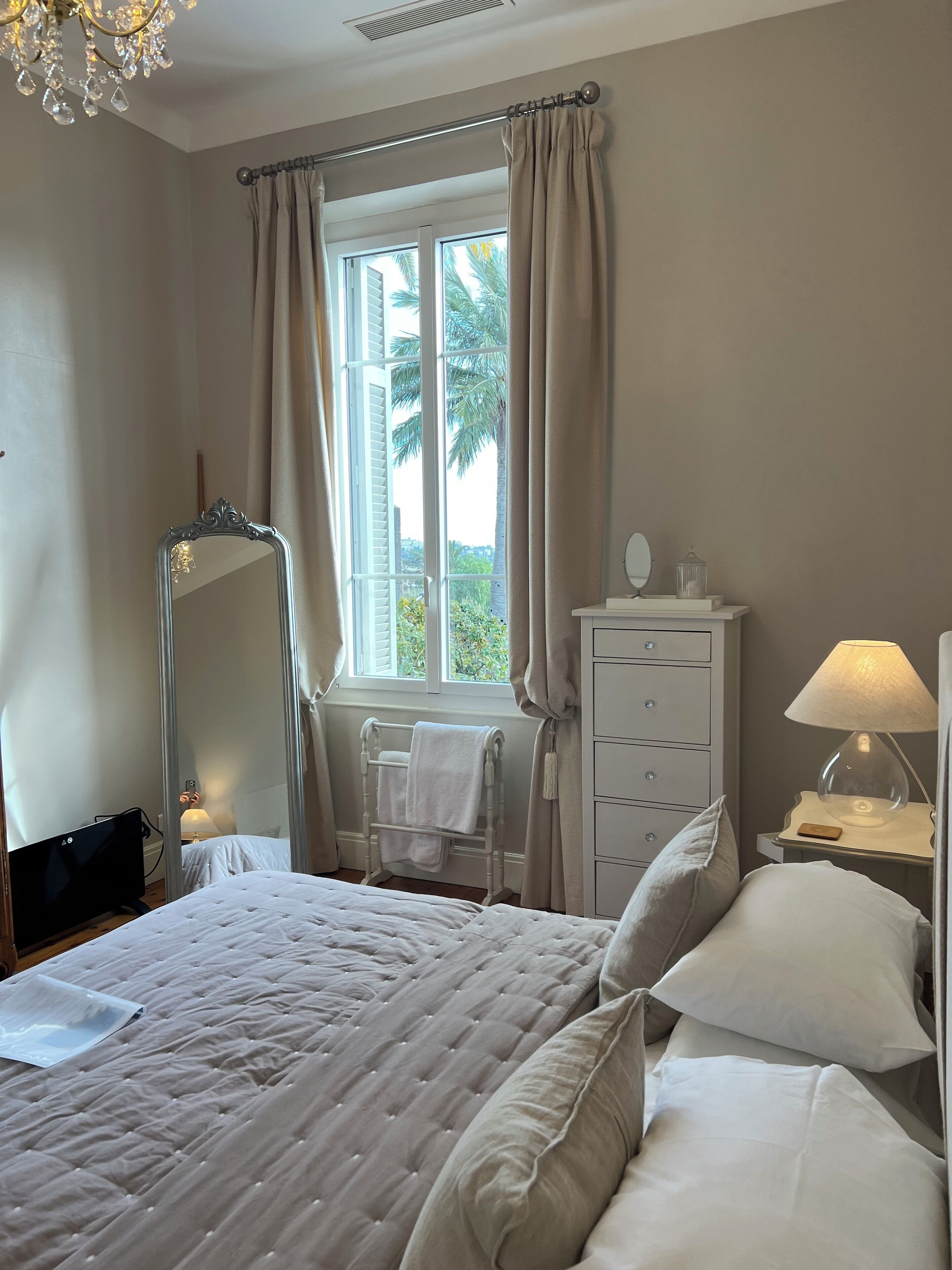Une chambre à coucher avec un lit, une table de chevet avec une lampe, une commode, une fenêtre avec des rideaux et un miroir.