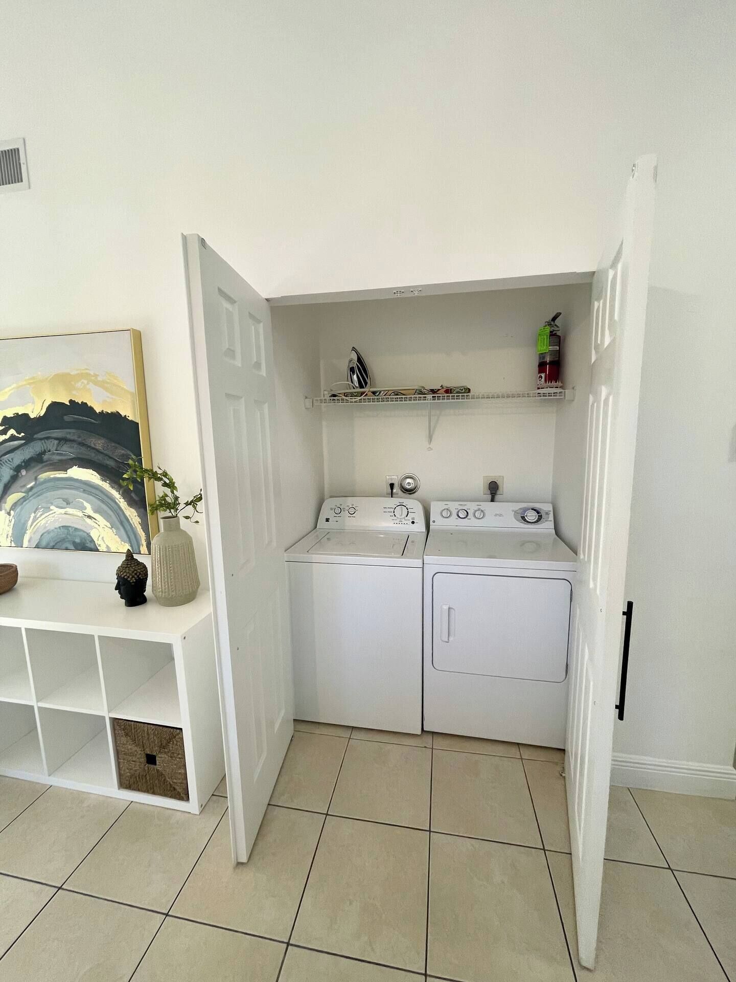 A laundry room with two washing machines, a drying machine, a shelf with cleaning supplies, and a decorative vase.