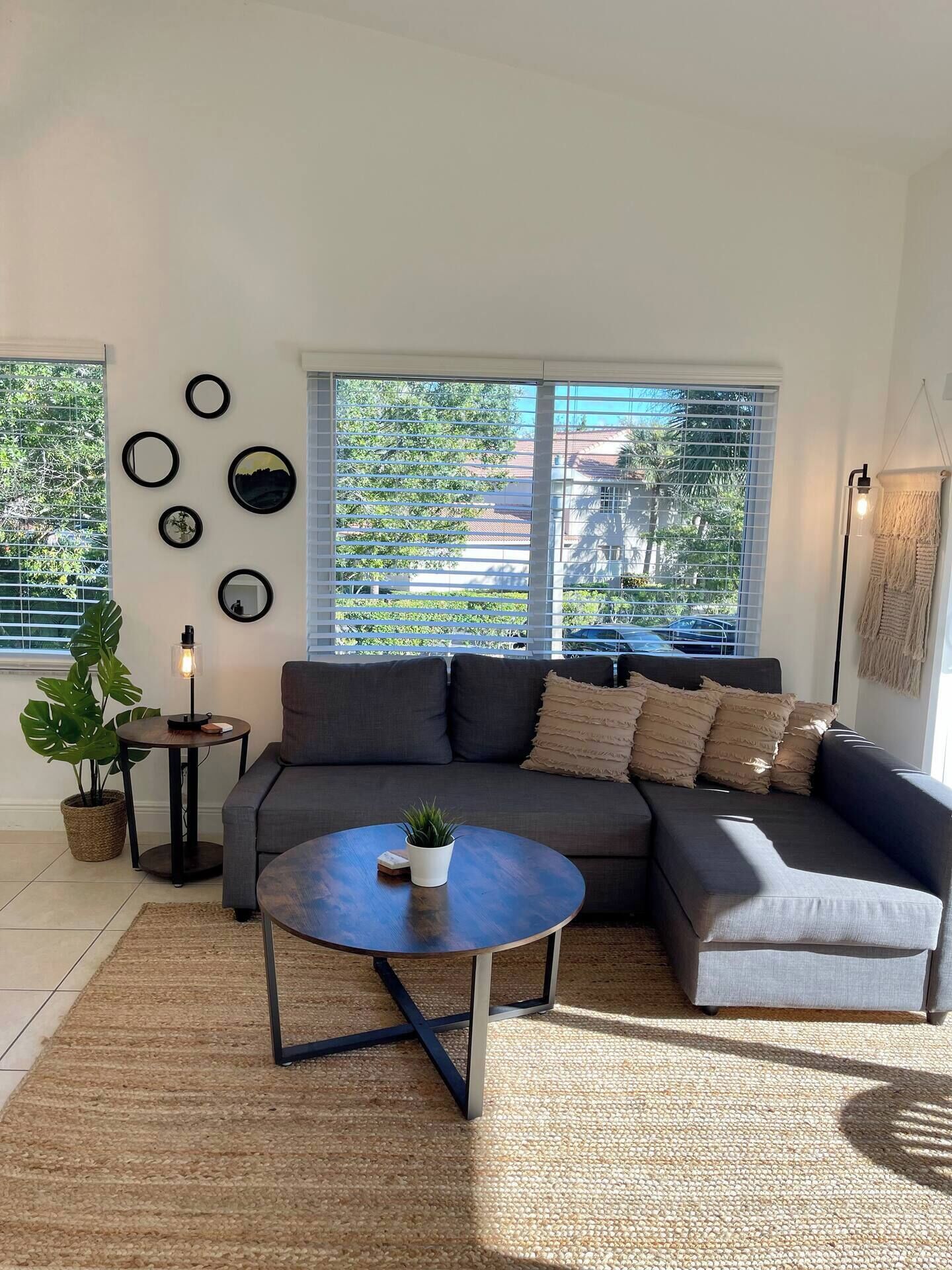 Ein modernes Wohnzimmer mit einem grauen Ecksofa, einem runden Couchtisch und einem großen Fenster mit Jalousien.