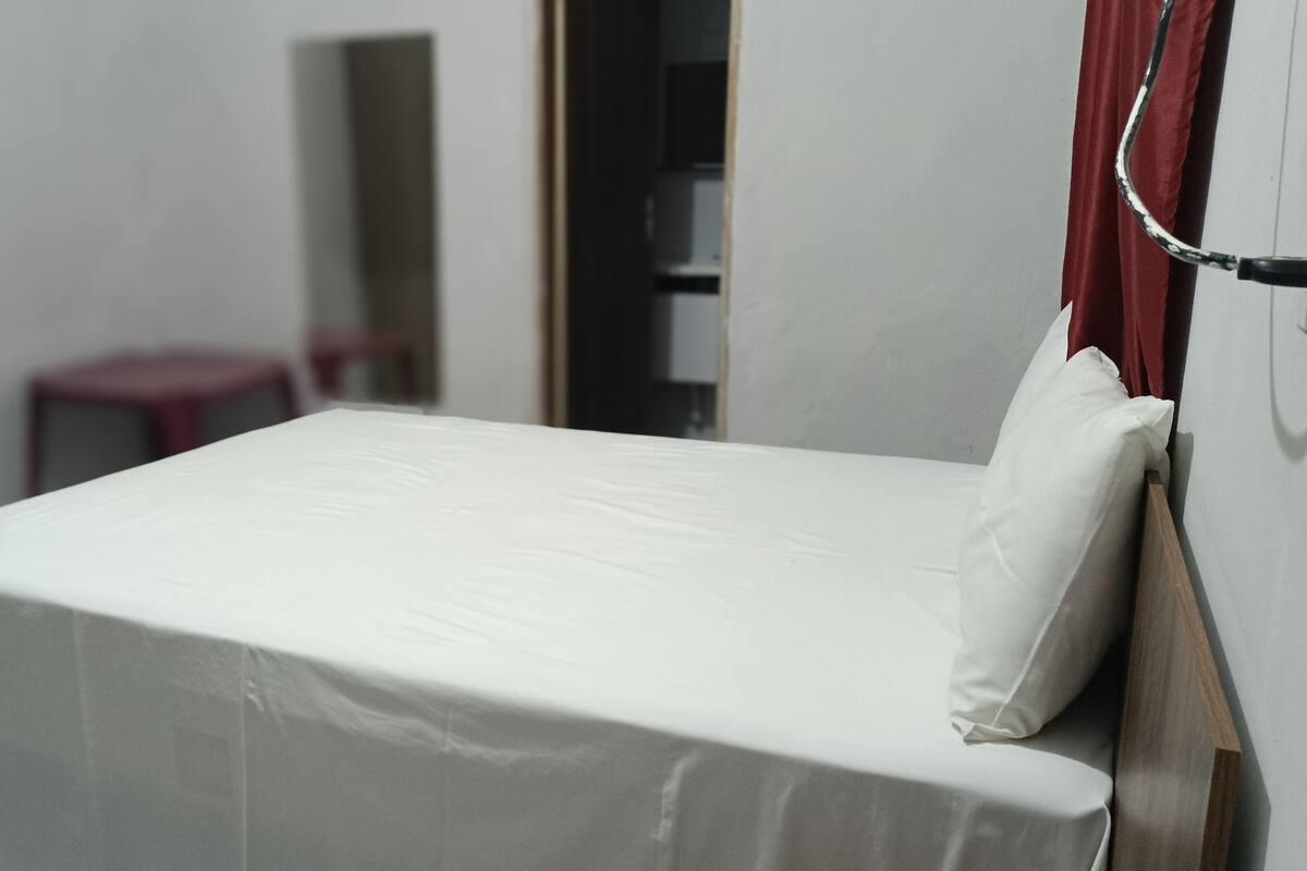 Ein kleines, ordentlich gemachtes Bett mit weißer Matratze und Kissen in einem Zimmer, das eine Holztür und einen roten Vorhang hat.