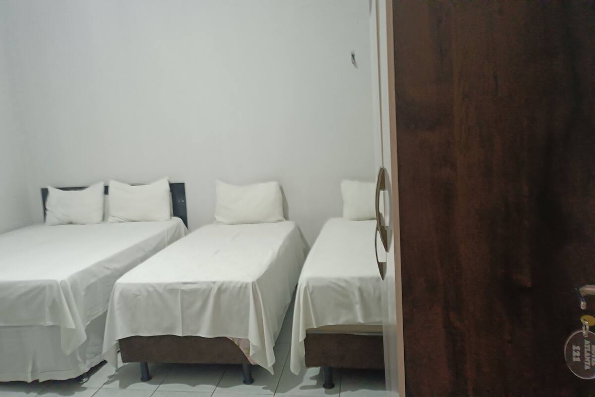 Ein Zimmer mit drei Einzelbetten, jedes mit weißer Bettwäsche und Kissen. Die Wände sind schlicht weiß, und es gibt eine Holztür.