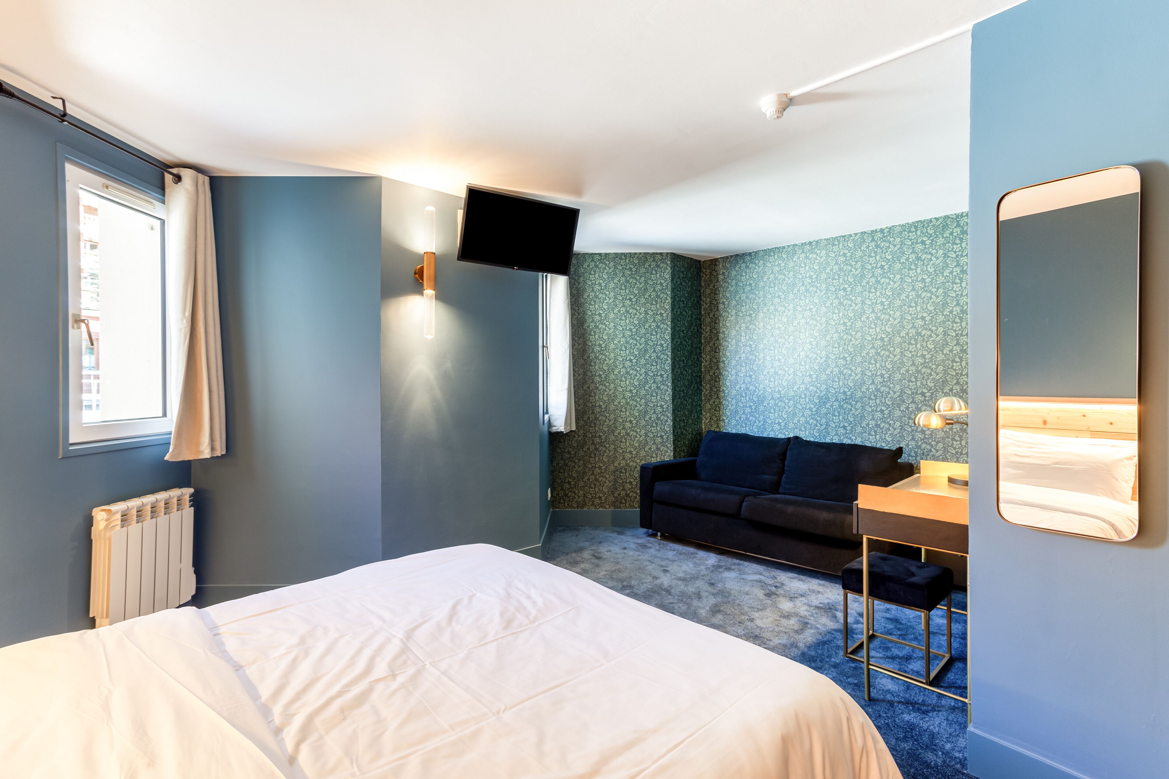 A hotel room with a bed, a desk, a chair, a sofa, a TV, and a window with curtains.