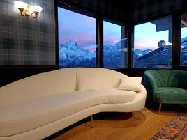A modern living room with a white sofa, a green armchair, and large windows offering a view of snow-covered mountains.