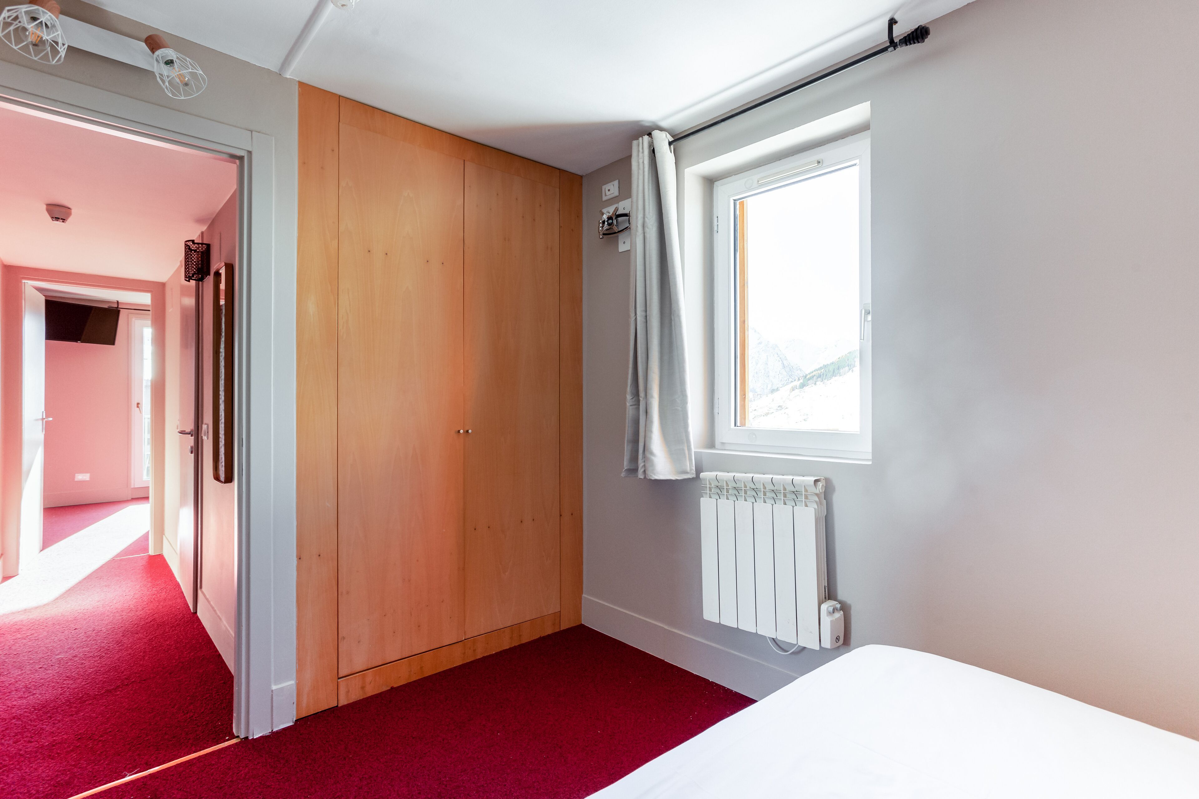 A room with a wooden wardrobe, a radiator, a window with curtains, and a red carpet.