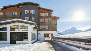 Un immeuble à plusieurs étages, doté d’une façade en bois et de grandes fenêtres, se dressant sur fond de montagnes enneigées.