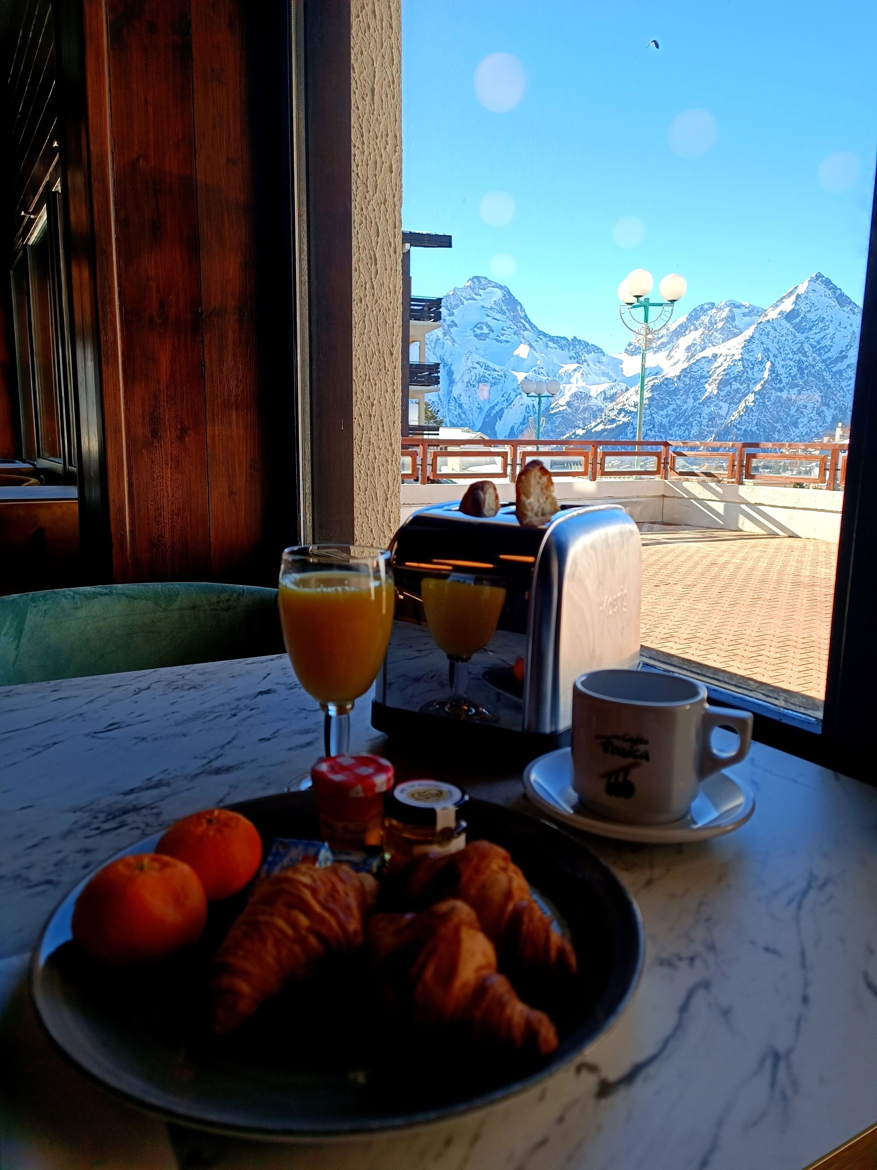 A breakfast table with croissants, oranges, and a coffee cup, overlooking a snowy mountain view.