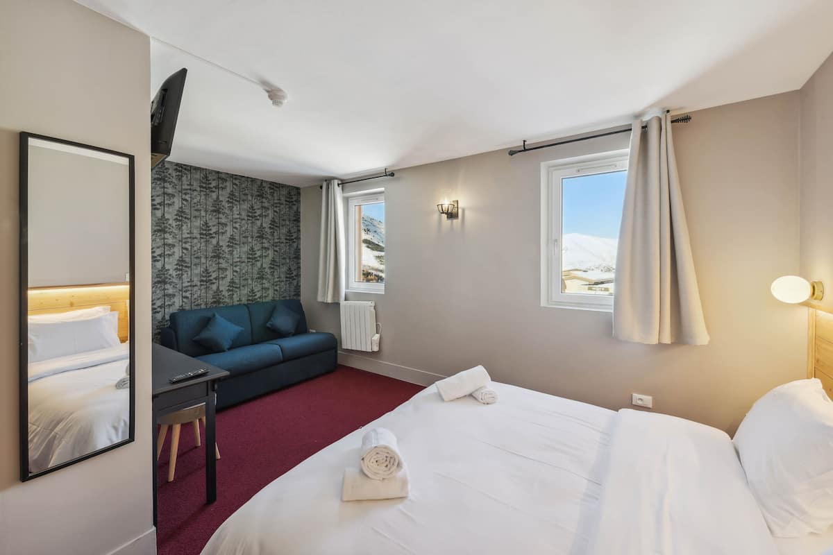 Ein Hotelzimmer mit Bett, Schreibtisch, Stuhl, Sofa, Spiegel, Wandfernseher, Fenster mit Vorhängen und Thermostat.