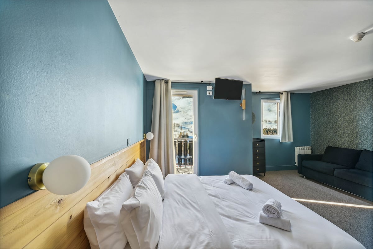Ein Hotelzimmer mit einem großen Bett, einem hölzernen Kopfteil, einem flachbildfernseher an der Wand, einem dunkelgrauen Sofa und Blick auf einen Balkon mit Sitzgelegenheit.