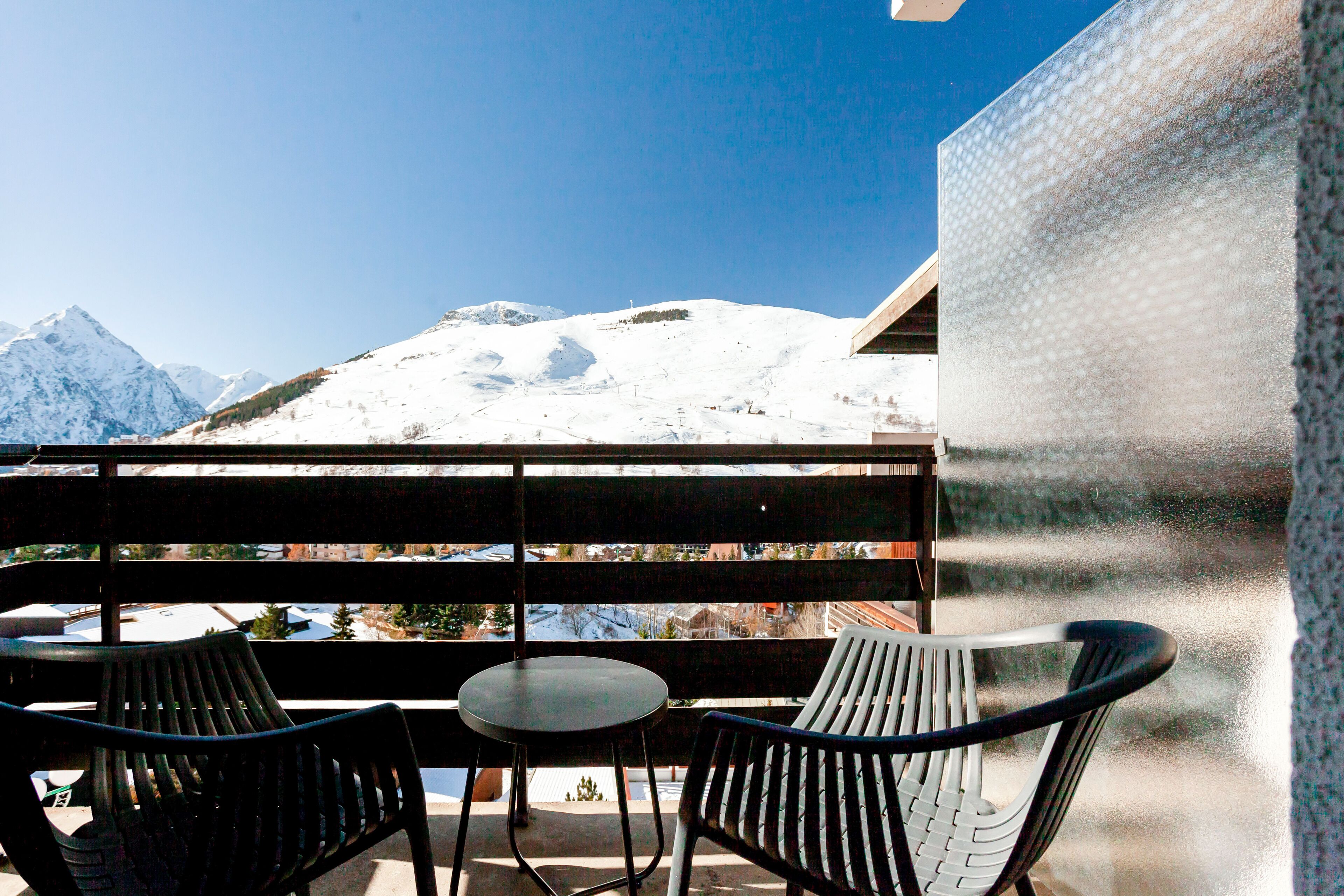 A balcony with two chairs and a table, overlooking a snowy mountain and a clear blue sky.