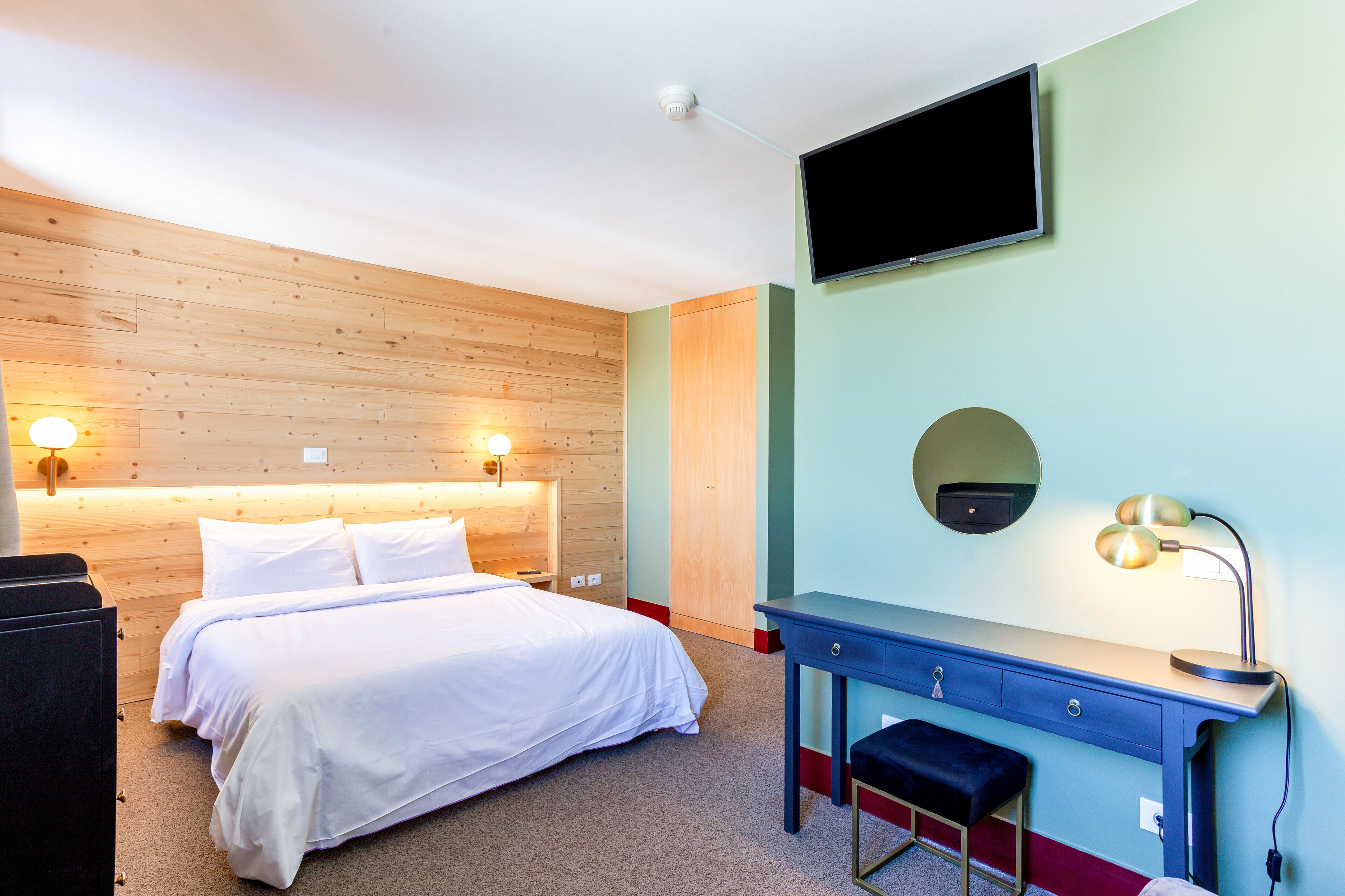 A hotel room with a wooden headboard, a bed with white linens, a desk with a chair, a television mounted on the wall, and a desk lamp.