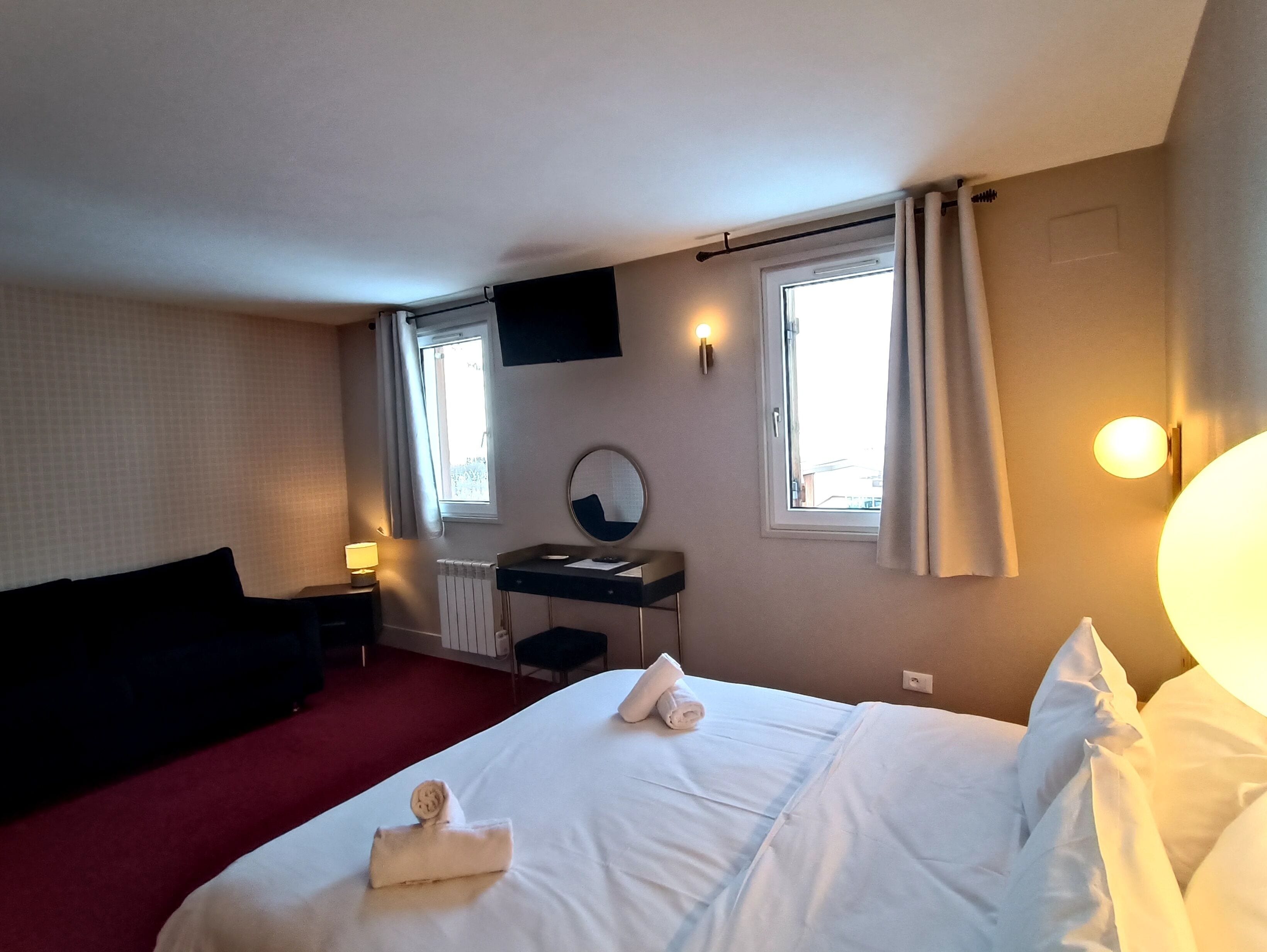 Ein Hotelzimmer mit einem Bett, einem Schreibtisch mit Spiegel, zwei Fenstern mit Vorhängen, einem an der Wand montierten Fernseher und einem roten Teppich.