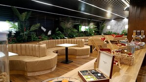 Un espace de détente moderne avec des sièges aux lignes courbes, un buffet proposant des plats et des boissons, et une variété de plantes.