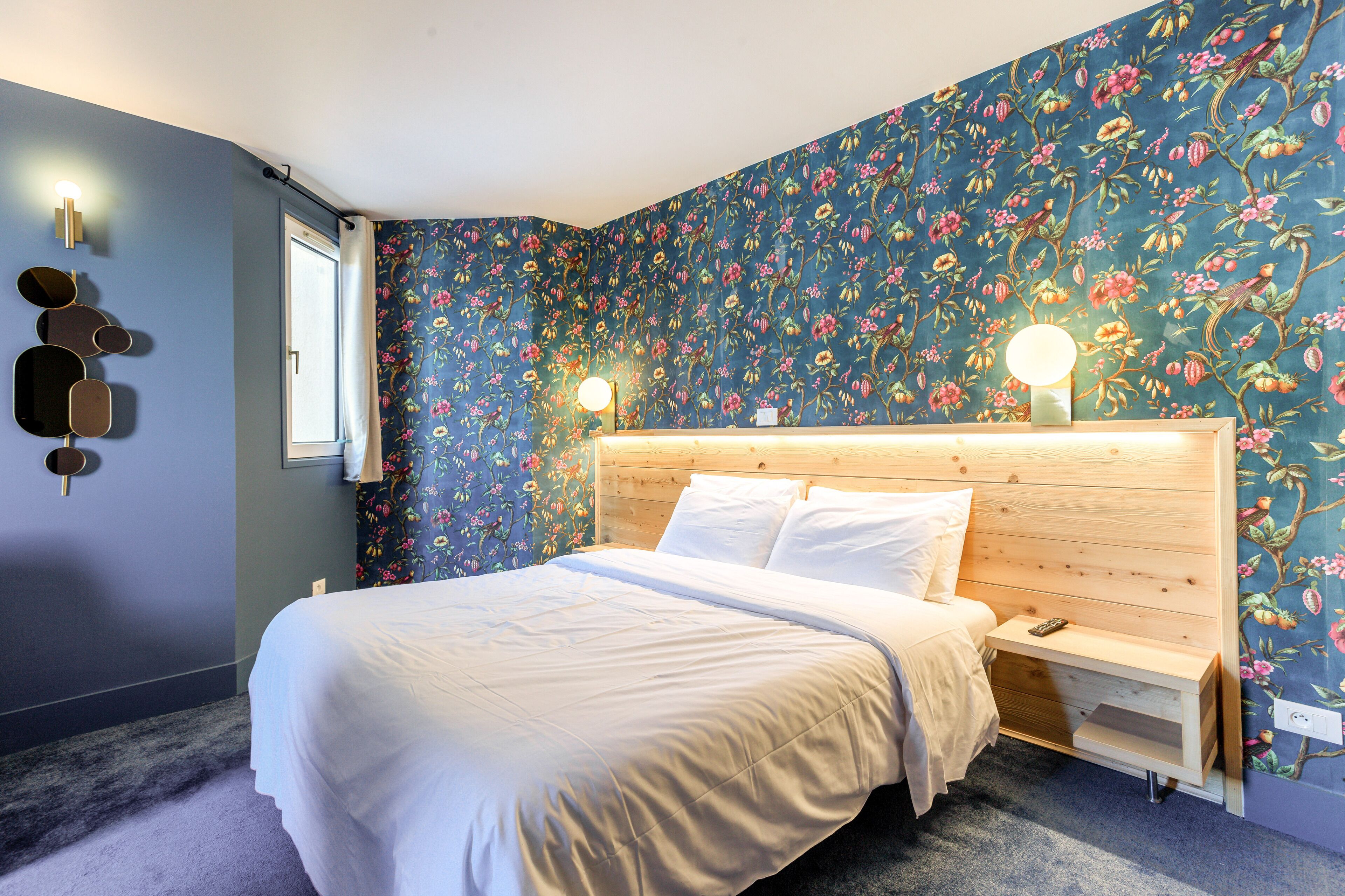 Un dormitorio con papel tapiz floral, cabecero de madera, mesita de noche con lámpara y ventana con cortinas.