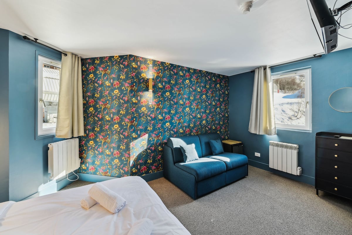 Ein Schlafzimmer mit blumigem Wandpapier, einem blauen Sofa, einem Bett, einem Schminktisch und einem Fenster mit Blick auf eine verschneite Landschaft.