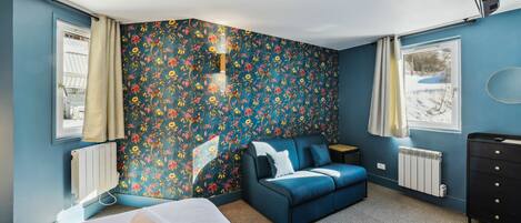 A bedroom with a floral wallpaper, a blue sofa, a bed, a dresser, and a window with a view of a snowy landscape.