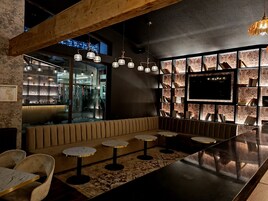 Un espace bar moderne avec un plafond en bois, un grand téléviseur mural et un coin salon doté de tables rondes et de chaises rembourrées.
