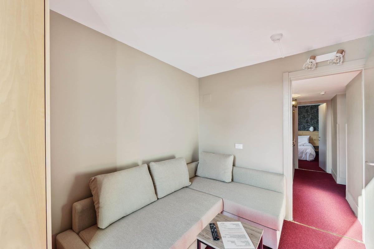Ein modernes Wohnzimmer mit einem beigefarbenen Ecksofa, einem kleinen Tisch mit Fernbedienung und einem rot ausgelegten Flur, der zum Schlafzimmer führt.