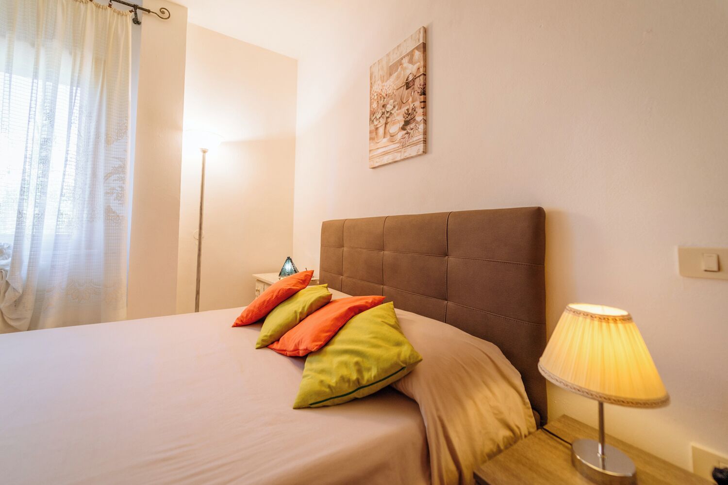 Cama bem arrumada com cabeceira marrom, luminária de mesa e almofadas coloridas.