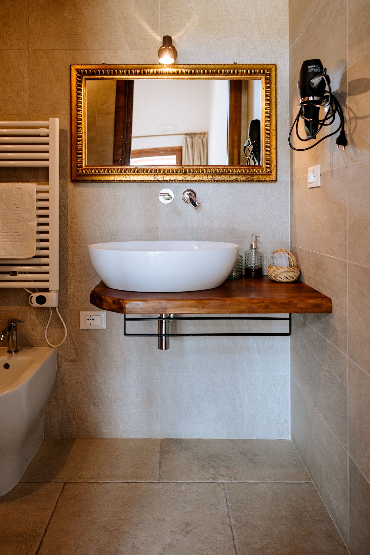 Un baño con un lavamanos blanco de diseño, un espejo con marco dorado, un perchero y un secador de pelo en la pared.