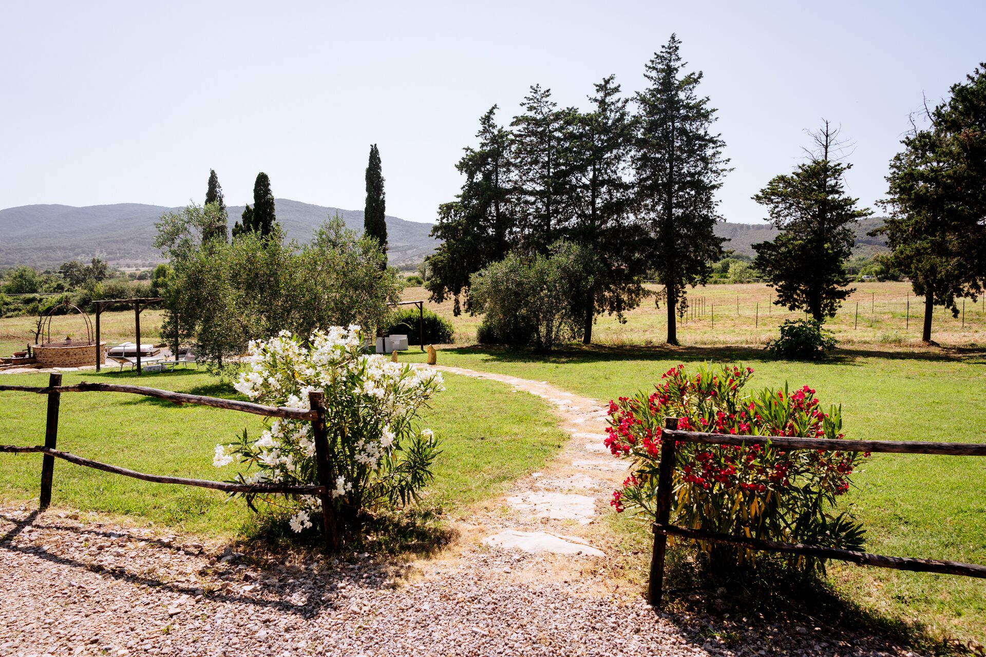 Ein Kiesweg führt durch einen von Zäunen umgebenen Garten mit weißen und roten Blüten, umgeben von Bäumen und einer Berglandschaft im Hintergrund.