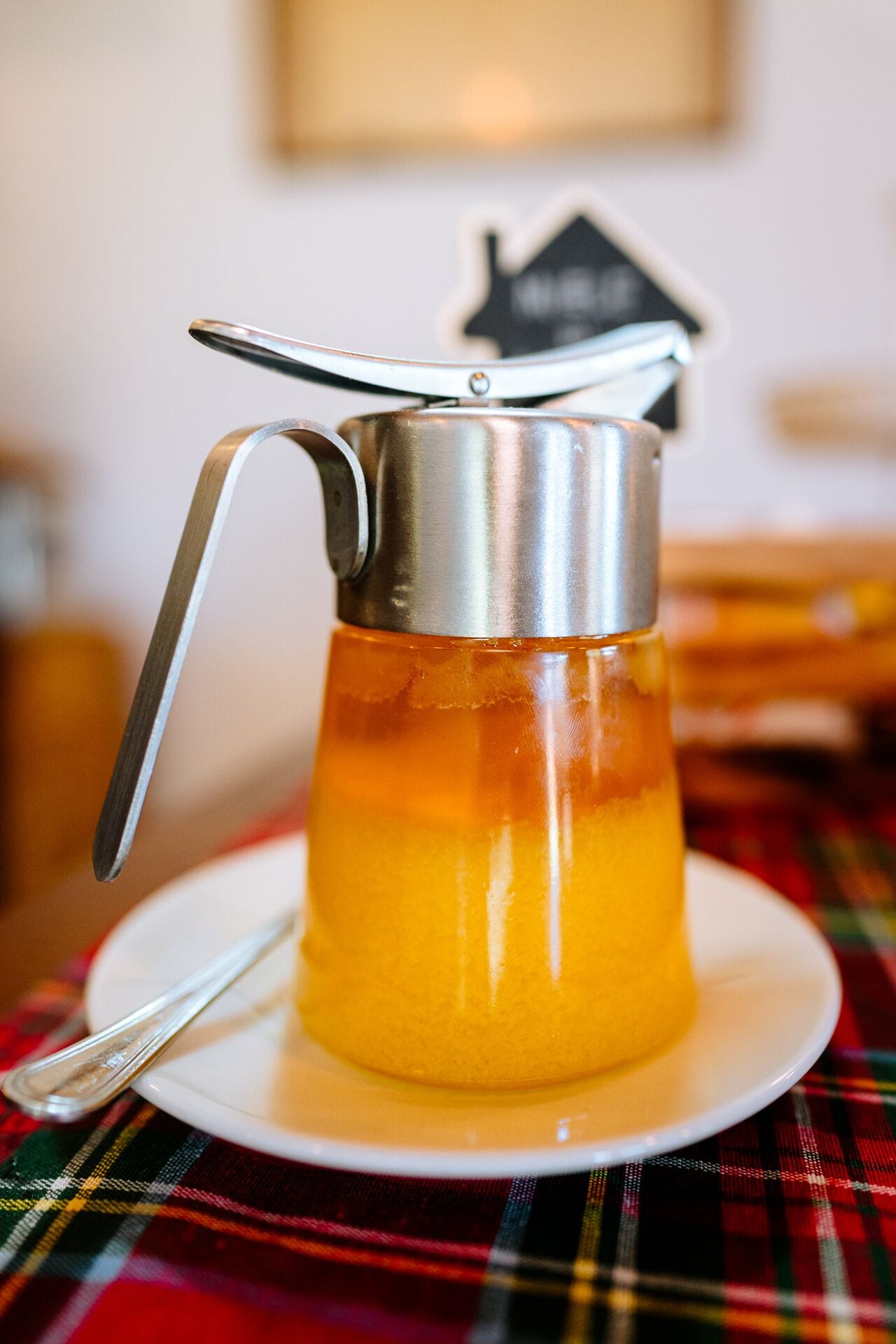 Una prensa francesa de metal con asa, llena de un líquido amarillo, sobre un plato blanco con una cuchara, colocada sobre un mantel escocés.