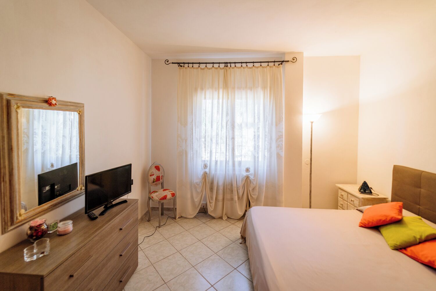 A bedroom with a bed, a TV, a chair, a dresser, a nightstand, and a window with curtains.